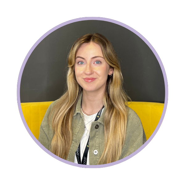 Woman with long blonde hair wearing a green jacket and white shirt, sitting on a yellow couch.
