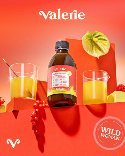 Bottle of Valerie daily essentials with a glass of juice on a red background, featuring the 'Wild Woman' label.